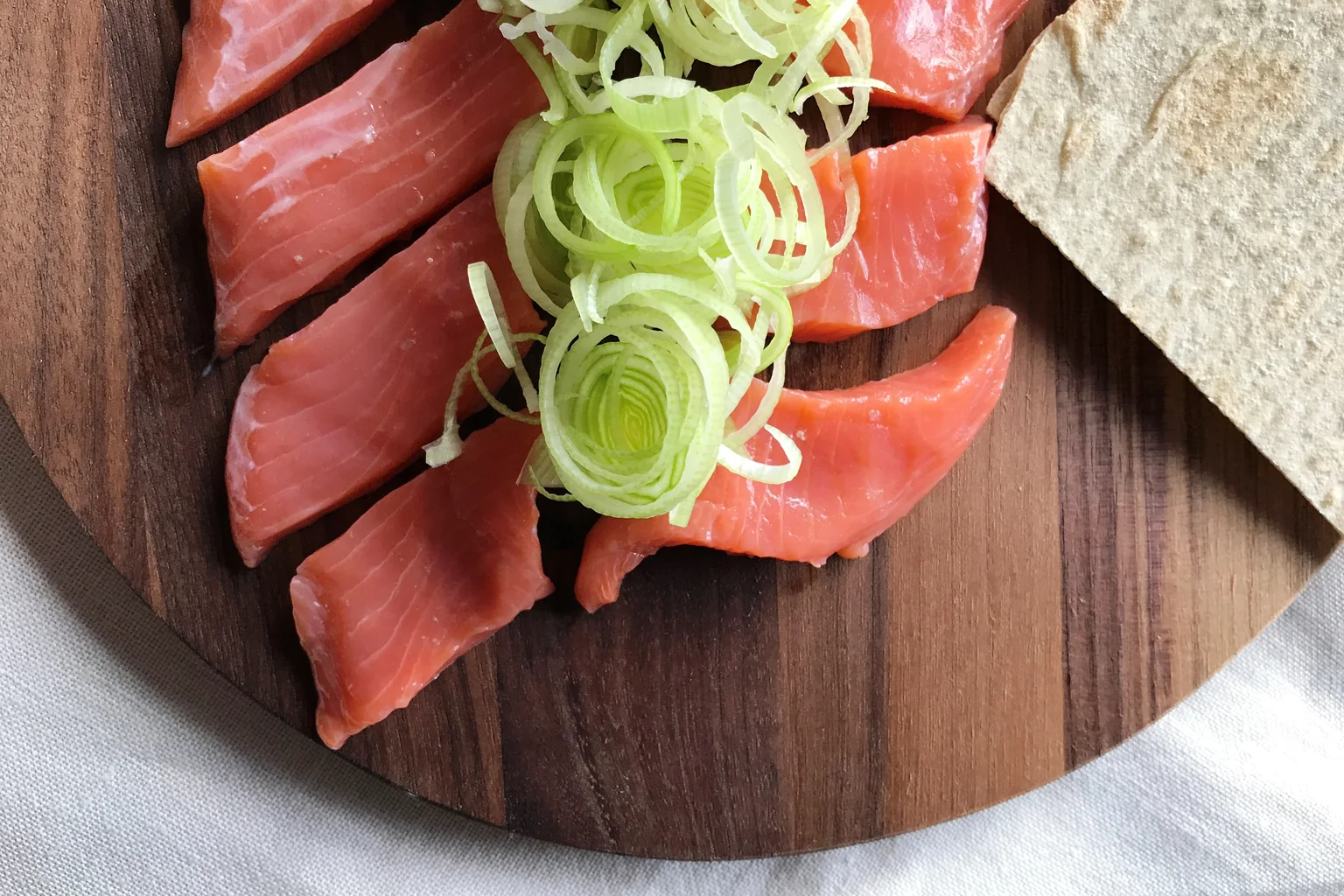 Skiver av rakfisk av ørret på rund trefjøl med strimlet purre og en potetlefse ved siden